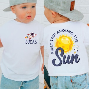 Op de afbeelding: Wit T-shirt met een afbeelding van een raket en een astronaut, de naam "LUCAS" staat eronder. De achterkant van het shirt heeft een zonafbeelding met de tekst "FIRST TRIP AROUND THE SUN". Het kind draagt een grijze baseballcap en een blauwe jeans.