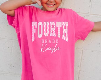 May include: A bright pink short-sleeved t-shirt featuring the word FOURTH in a white patterned block font, with the word GRADE in a smaller white font below it, and the name Kayla written in a white cursive script at the bottom center.