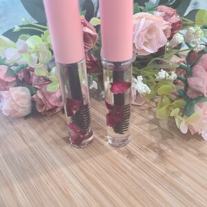 May include: Two clear glass bottles with pink caps filled with a clear liquid and dried rose petals. The bottles have a black mascara wand inside.