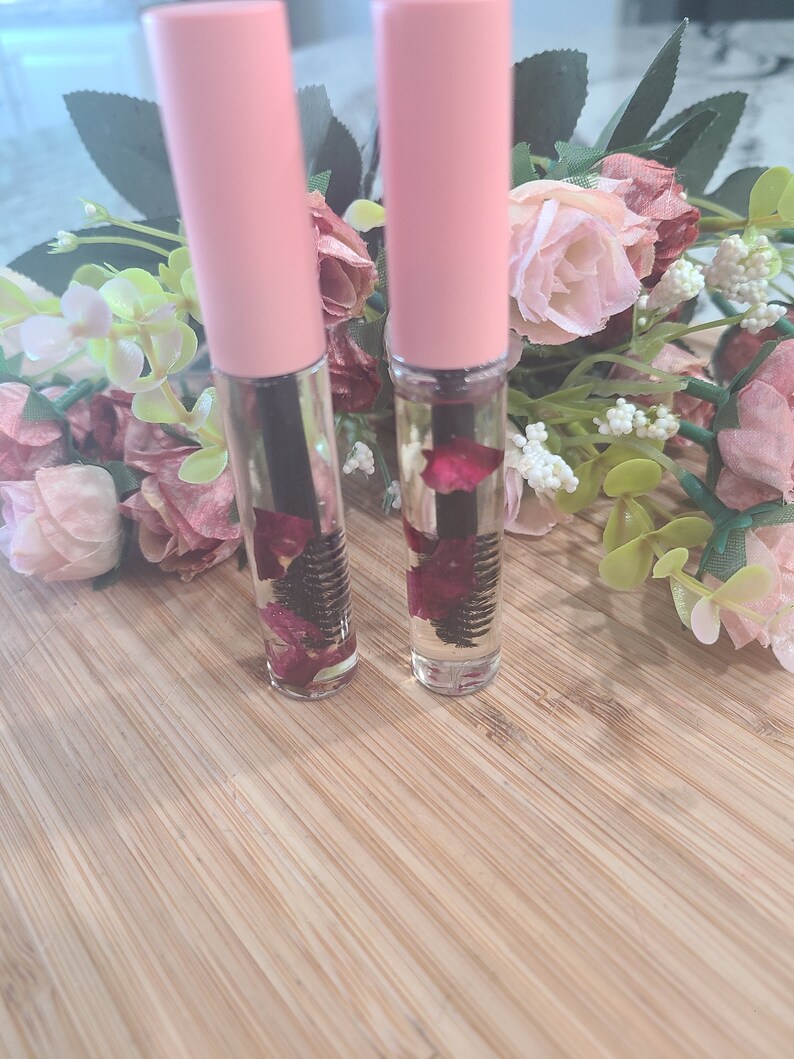 May include: Two clear glass bottles with pink caps filled with a clear liquid and dried rose petals. The bottles have a black mascara wand inside.