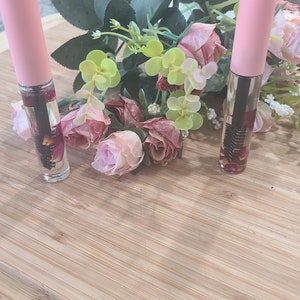 May include: Two pink bottles with clear liquid and dried flowers inside, sitting on a light brown wooden surface with pink flowers in the background.