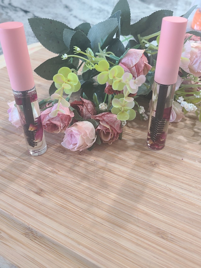May include: Two pink bottles with clear liquid and dried flowers inside, sitting on a light brown wooden surface with pink flowers in the background.
