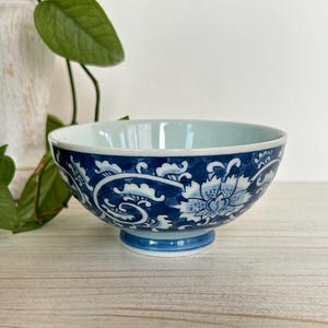 Puede incluir: Un cuenco de cerámica con un diseño floral azul y blanco. El cuenco tiene un interior azul claro y un exterior azul oscuro con patrones florales blancos. Se apoya sobre una pequeña base redonda y se exhibe sobre una superficie de madera clara.