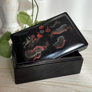 Puede incluir: Caja rectangular negra con tapa con bisagras, con un diseño decorativo de dragones en rojo, gris y blanco. La caja está abierta, revelando el interior. La caja está sobre una superficie de madera clara.