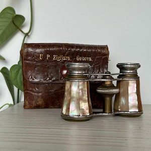 May include: Antique opera glasses with mother-of-pearl inlays and silver accents. The glasses are next to a brown leather case embossed with "U.P. Zigliara, Genova". The case has a crocodile skin texture. The opera glasses are on a light-colored surface.