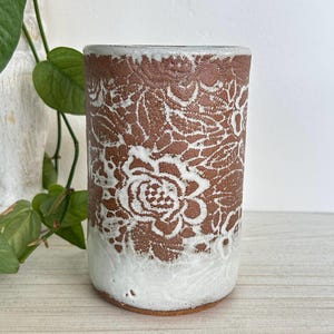 Jarrón cilíndrico vintage con relieve de encaje floral, cerámica de gres estilo bohemio, tonos tierra y esmalte blanco.