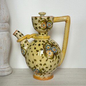 May include: A ceramic pitcher with a bulbous body, a small spout, and a large handle. The pitcher is painted with floral designs in blue, orange, and green on a yellow background. The handle and spout are yellow with a rope-like design.