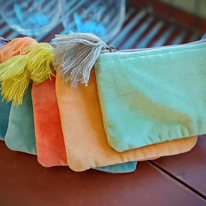 May include: Five colorful velvet pouches with tassels. The pouches are stacked on top of each other, with the lightest color on top and the darkest color on the bottom. The colors are light blue, teal, orange, peach, and purple.