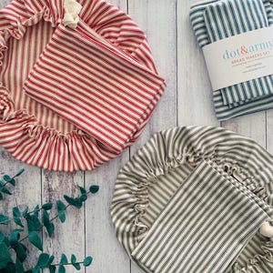 Könnte beinhalten: Ein Set für Brotbackwaren, darunter zwei gestreifte Schüsselbezüge und eine passende Tasche in Rot und Weiß sowie Grau und Weiß. Ein gefaltetes Set aus blau-weiß gestreiften Brottüchern ist ebenfalls enthalten. Das Etikett lautet "dot & army BREAD MAKERS SET".