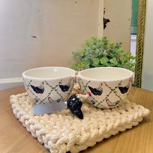 May include: Two white ceramic bowls with a chicken pattern, resting on a cream-colored, hand-knitted coaster. A chicken-shaped cheese knife sits between the bowls. The bowls and knife are on a wooden table.