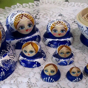 May include: A collection of Russian nesting dolls, or Matryoshka dolls, in various sizes. The dolls feature a blue and white floral design with a painted face. The largest doll is approximately 6 inches tall.