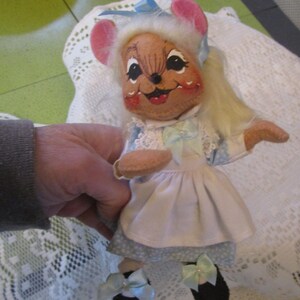 May include: A vintage mouse doll with a painted face, pink ears, and blonde hair. The doll wears a blue and white dress with a white apron and black shoes with blue bows. The doll is being held by a person.