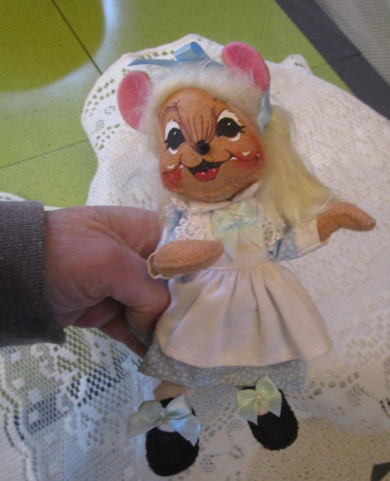 May include: A vintage mouse doll with a painted face, pink ears, and blonde hair. The doll wears a blue and white dress with a white apron and black shoes with blue bows. The doll is being held by a person.