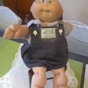 May include: A vintage Cabbage Patch Kids doll with a round face, blue eyes, and a tuft of blonde hair. The doll wears a denim overall with a gingham shirt underneath. The doll is seated on a small wooden chair.