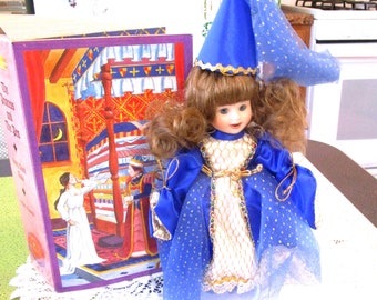 Puede incluir: Una muñeca de porcelana vestida con un vestido de princesa azul y un sombrero a juego con un velo transparente. La muñeca tiene el pelo rizado castaño y está colocada junto a un libro infantil titulado "La Princesa y el Guisante".