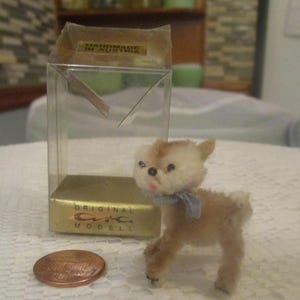 May include: A miniature deer figurine with a light brown body, a white head, and a blue bow. The figurine is next to a clear plastic box with a gold base and the text "ORIGINAL CVC MODELL". A penny is also in the image.