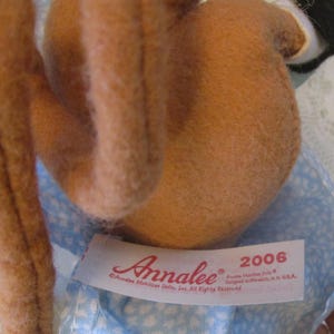 May include: A close-up of a doll with brown fur and a blue patterned dress. The doll has a tag that reads "Annalee" and "2006". The doll is a collectible item.