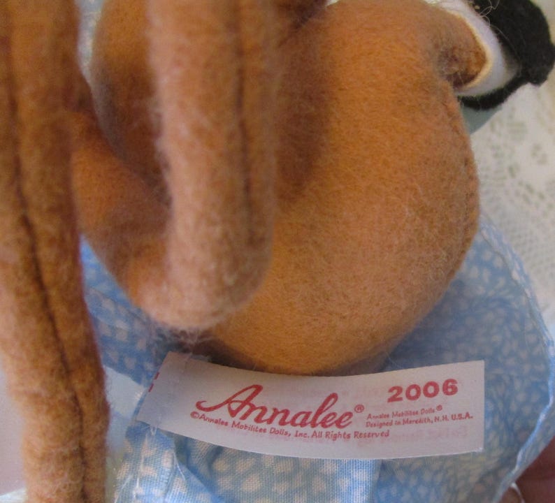 May include: A close-up of a doll with brown fur and a blue patterned dress. The doll has a tag that reads "Annalee" and "2006". The doll is a collectible item.