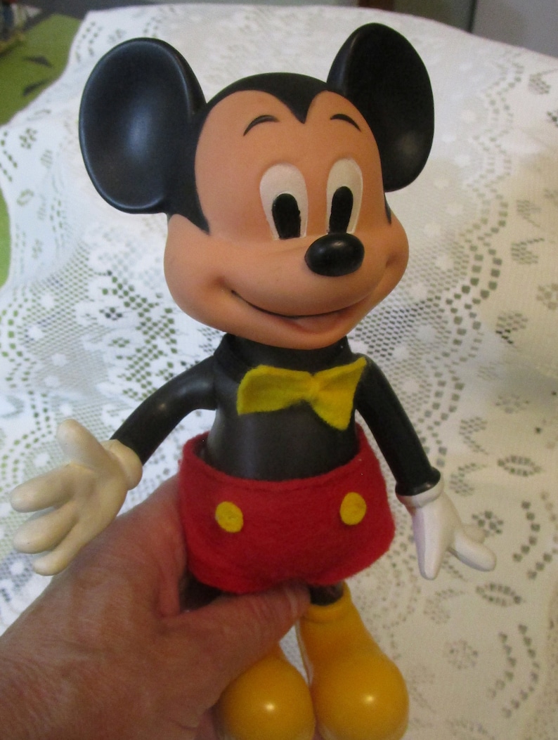 May include: A vintage Mickey Mouse figurine. The figure has a black body, large black ears, and a tan face with a smiling expression. He wears red shorts with yellow buttons, a yellow bow tie, and yellow shoes. His hands are white.
