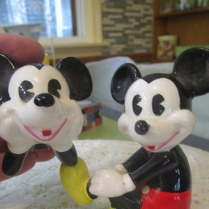 May include: A pair of ceramic Mickey Mouse figurines. The figurine on the left is holding its chin and has a yellow shoe. The figurine on the right is standing with its right hand raised and is wearing red shorts.
