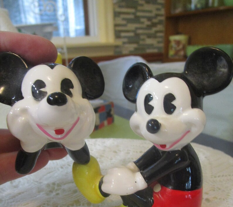 May include: A pair of ceramic Mickey Mouse figurines. The figurine on the left is holding its chin and has a yellow shoe. The figurine on the right is standing with its right hand raised and is wearing red shorts.