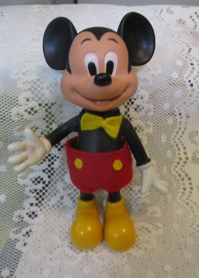 May include: A Mickey Mouse figurine with a smiling face, black ears, and a yellow bow tie. The figure wears red shorts with yellow buttons, black arms, white gloves, and yellow shoes. The toy is a classic Disney character.