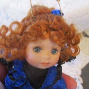 May include: A porcelain doll with curly red hair, blue eyes, and a blue and black dress. The doll is wearing two gold hair clips.