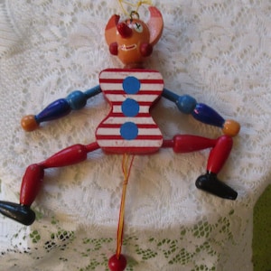 May include: A vintage wooden pull-string toy clown with articulated limbs. The clown has a red and white striped torso, blue arms, and red legs. The head is orange with red ears and a red nose. The toy is suspended by a yellow string.