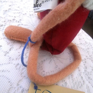 K&ouml;nnte beinhalten: Handgefertigte Puppe mit rotem Stoffk&ouml;rper, hellbraunen Gliedma&szlig;en und einer wei&szlig;en Strickm&uuml;tze. Die Puppe hat ein Etikett mit der Aufschrift "Carlee&reg; 2011". Ein blauer Faden ist am Bein der Puppe befestigt und ein beiges Etikett.