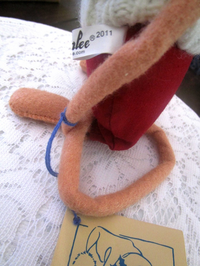 K&ouml;nnte beinhalten: Handgefertigte Puppe mit rotem Stoffk&ouml;rper, hellbraunen Gliedma&szlig;en und einer wei&szlig;en Strickm&uuml;tze. Die Puppe hat ein Etikett mit der Aufschrift "Carlee&reg; 2011". Ein blauer Faden ist am Bein der Puppe befestigt und ein beiges Etikett.