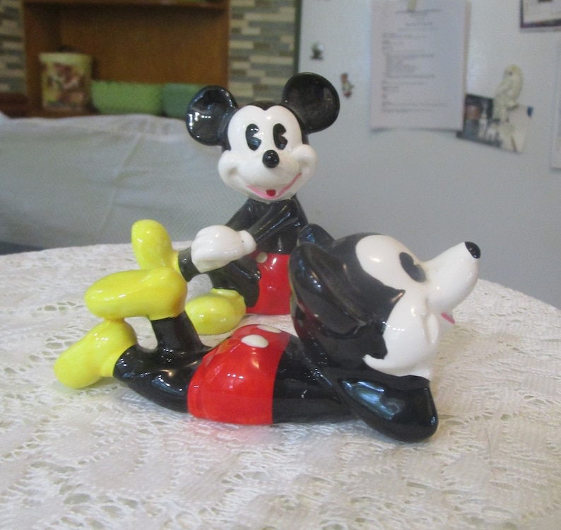 May include: Two ceramic Mickey Mouse figurines. One is standing with his arms crossed and the other is lying down with his head turned to the side. Both figurines are painted in black, white, red, and yellow.