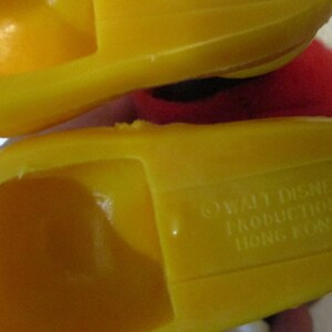 May include: Close-up of two yellow plastic toy shoes. The underside of one shoe is visible, with the text "&copy; WALT DISNEY PRODUCTION HONG KONG" embossed. The shoes are a bright, solid yellow colour.