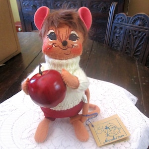 May include: A vintage Analee doll in the shape of a mouse, holding a large red apple. The doll has a white sweater, red shorts, and a brown fur-like hair. A tag with the words "Analee Dolls" is attached.