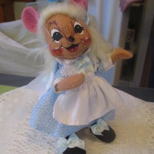 May include: A handmade mouse doll wearing a light blue dress with white polka dots, a white apron, and black shoes with blue bows. The mouse has pink ears, a brown face, and white hair. The doll is posed on a white lace surface.