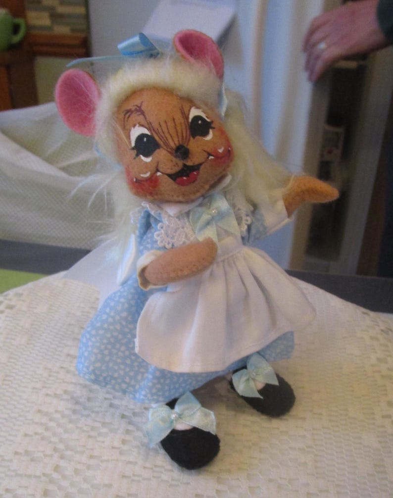 May include: A handmade mouse doll wearing a light blue dress with white polka dots, a white apron, and black shoes with blue bows. The mouse has pink ears, a brown face, and white hair. The doll is posed on a white lace surface.