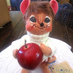 K&ouml;nnte beinhalten: Eine Vintage-Annalee-Puppe, eine Maus mit bemaltem Gesicht, tr&auml;gt einen wei&szlig;en Pullover und h&auml;lt einen roten Apfel. Die Puppe hat braunes Fell und rosa Ohren. Ein Etikett mit dem Annalee Dolls-Logo ist sichtbar.