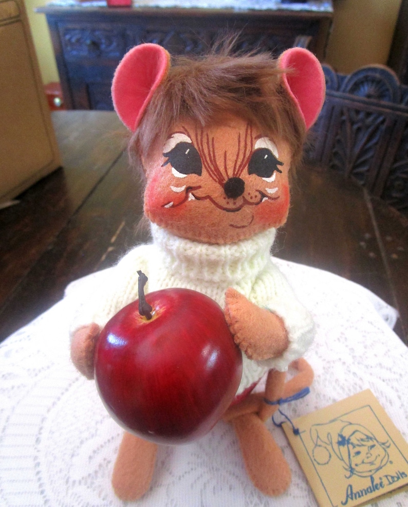 K&ouml;nnte beinhalten: Eine Vintage-Annalee-Puppe, eine Maus mit bemaltem Gesicht, tr&auml;gt einen wei&szlig;en Pullover und h&auml;lt einen roten Apfel. Die Puppe hat braunes Fell und rosa Ohren. Ein Etikett mit dem Annalee Dolls-Logo ist sichtbar.