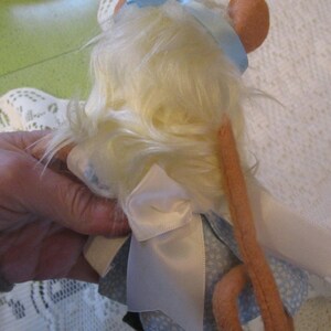 May include: A handmade doll of a mouse with light yellow fur, wearing a blue dress with white floral patterns and a white apron. The mouse has pink ears and a long, pink tail. A light blue ribbon is tied on its head.