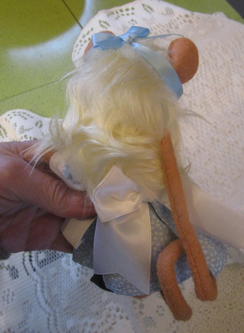 May include: A handmade doll of a mouse with light yellow fur, wearing a blue dress with white floral patterns and a white apron. The mouse has pink ears and a long, pink tail. A light blue ribbon is tied on its head.