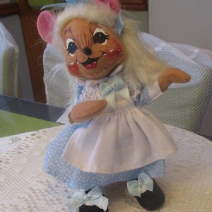 May include: A handmade doll in the shape of a mouse, wearing a blue and white dress with a white apron. The doll has pink ears, a smiling face, and black shoes with blue bows. The doll has blonde hair and a blue bow on its head.