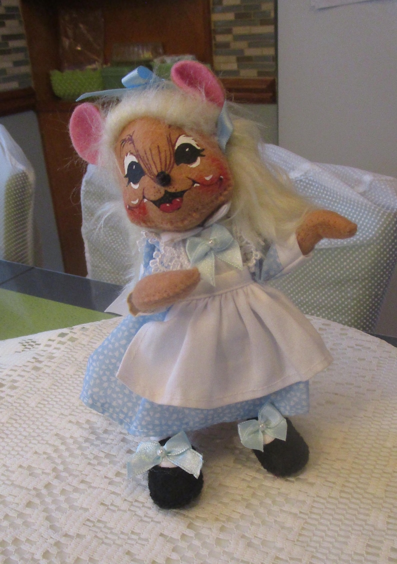 May include: A handmade doll in the shape of a mouse, wearing a blue and white dress with a white apron. The doll has pink ears, a smiling face, and black shoes with blue bows. The doll has blonde hair and a blue bow on its head.