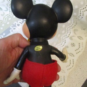 May include: A vintage Mickey Mouse figure, seen from the rear. The figure has a black head with large ears, a black body, red shorts, yellow shoes, and white gloves. A gold sticker on the back reads "MADE IN HONG KONG."