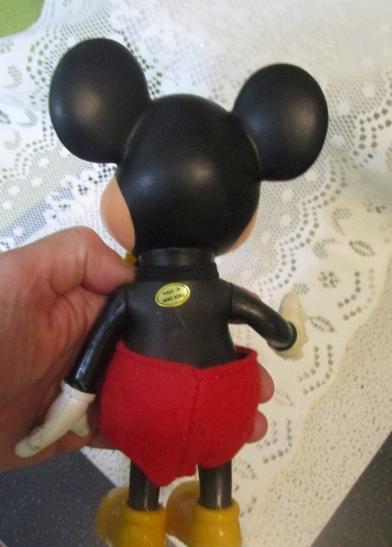 May include: A vintage Mickey Mouse figure, seen from the rear. The figure has a black head with large ears, a black body, red shorts, yellow shoes, and white gloves. A gold sticker on the back reads "MADE IN HONG KONG."