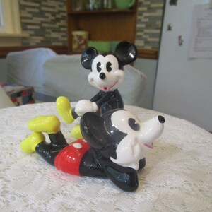 May include: Two ceramic Mickey Mouse figurines. One is standing with a yellow shoe and the other is lying down with a red and white shirt.