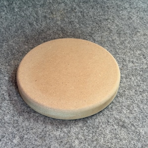 Könnte beinhalten: Ein kreisförmiges Objekt mit flacher Oberseite, einer glatten, hellbraunen Oberfläche und einer abgerundeten Kante. Der Gegenstand weist eine gleichmäßige, matte Textur und eine durchgehend beige Farbe auf, was eine einfache geometrische Form ergibt.