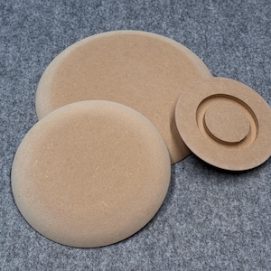 Op de afbeelding: Een verzameling van drie onafgewerkte, lichtbruine, cirkelvormige houten stukken. De grootste is een massieve schijf, de volgende is een iets kleinere schijf en de kleinste heeft een verhoogde, concentrische ring in het midden. Op een grijze ondergrond.