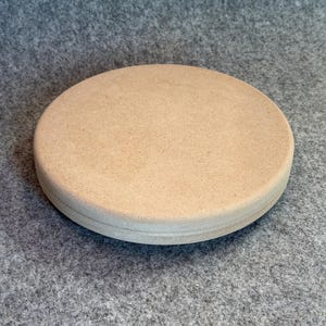 Könnte beinhalten: Ein runder, flacher Präsentationsständer aus Holz mit einer glatten, hellbraunen und unbehandelten Oberfläche. Das Objekt weist eine deutliche horizontale Rille entlang der Seitenkante auf, die dieser runden Holzbasis ein geschichtetes Aussehen verleiht.