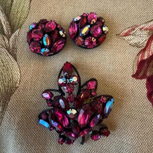 May include: A set of vintage jewelry featuring two round clip-on earrings and a leaf-shaped brooch. The pieces are adorned with vibrant, fuchsia-colored rhinestones and iridescent accents, set against a black metal frame. The jewelry is displayed on a patterned fabric.