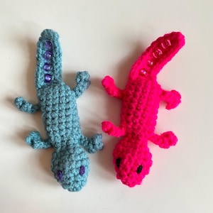 May include: Two handmade crochet lizards. One is light blue with purple bead accents and the other is bright pink with clear bead accents. Both have small arms and legs and black eyes.