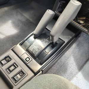 May include: Close-up of a car's interior showing a black gear shifter with a silver lever and a gray carpet. The gear shifter has a black plastic cover with the letters "P, R, N, D, 2, L" printed on it. There are three buttons below the gear shifter labeled "UP,  DOWN, and  PWR".